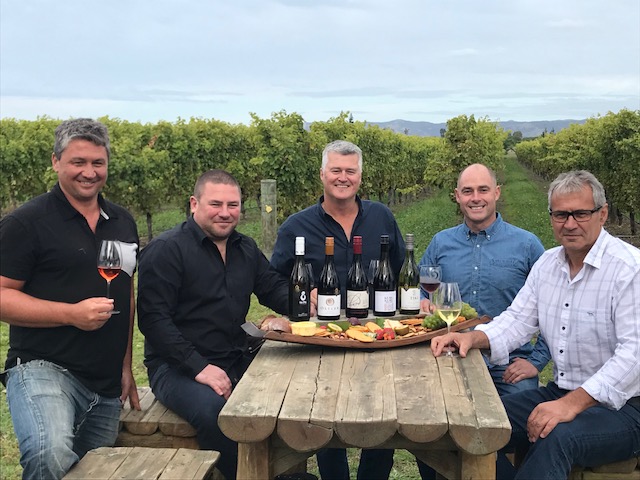 Five men sitting around a table with five bottles of wine and a platter of food being presented