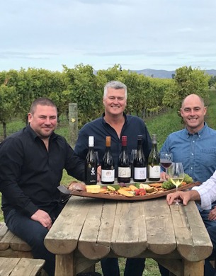 Five men sitting around a table with five bottles of wine and a platter of food being presented