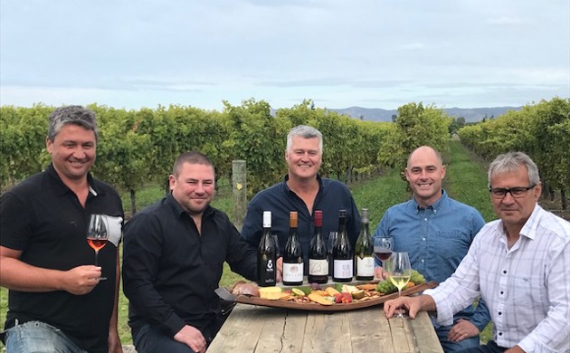 Five men sitting around a table with five bottles of wine and a platter of food being presented