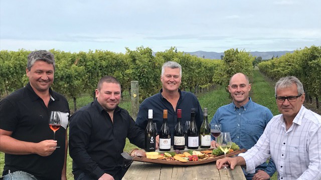 Five men sitting around a table with five bottles of wine and a platter of food being presented