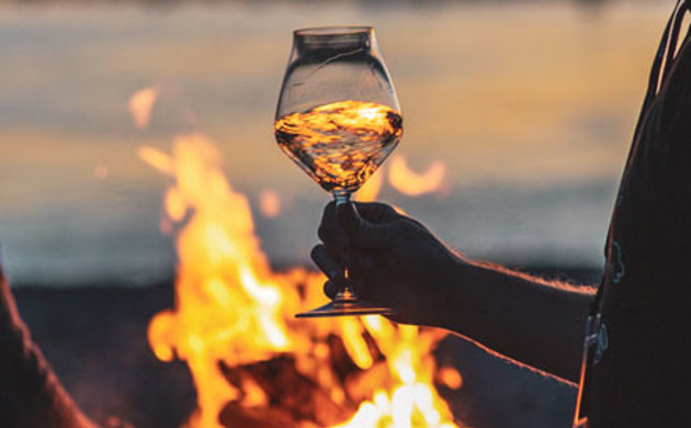 A glass of Pinot Gris being held by a person infront of an outdoor fire.