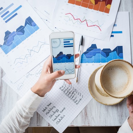 A person holding coffee while looking at stats on a smartphone and documents
