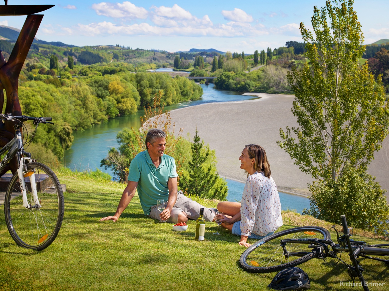 A couple sitting on a grassy knoll drinking wine with a bowl of grapes next to two bikes.